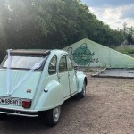 Une 2 CV vert pale devant la banderole des jardins du temps
